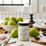 Candle jar labeled 'Apple Martini' on a table with apples, pears, and cinnamon sticks.