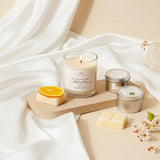 Candle and small containers on a wooden board with white fabric and orange slice in the background