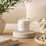 One Love soy candle by Nurse Calm Aromatherapy™ displayed in a cozy neutral-toned living space with soft blankets, natural textures, and warm ambient lighting. Clear glass candle with lit flame featuring tropical citrus, pineapple, coconut, rum, and sugar fragrance notes.
