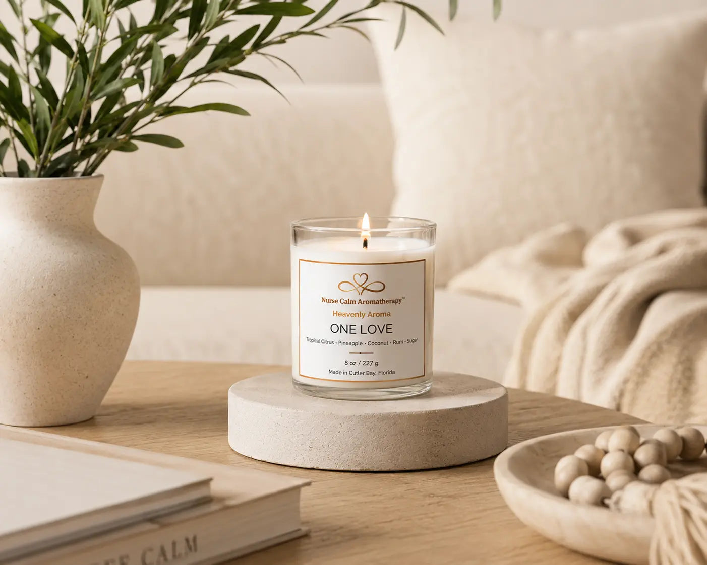 One Love soy candle by Nurse Calm Aromatherapy™ displayed in a cozy neutral-toned living space with soft blankets, natural textures, and warm ambient lighting. Clear glass candle with lit flame featuring tropical citrus, pineapple, coconut, rum, and sugar fragrance notes.
