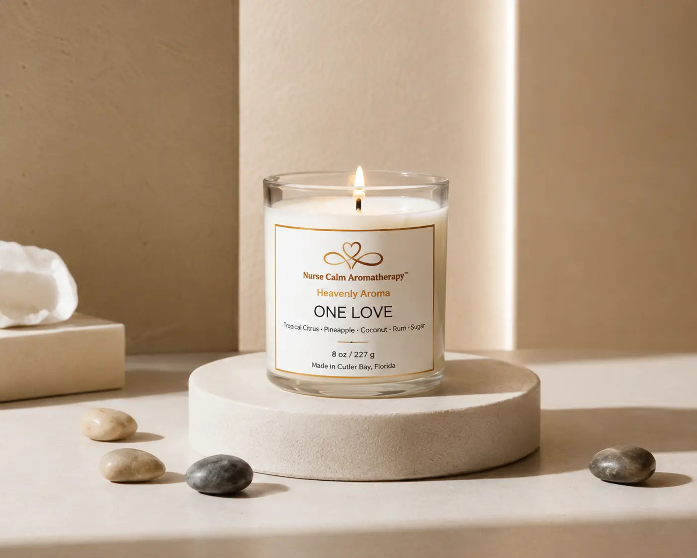 One Love soy candle by Nurse Calm Aromatherapy™ displayed on a neutral cream pedestal with soft spa-inspired décor and warm natural lighting. Clear glass candle with lit flame featuring tropical citrus, pineapple, coconut, rum, and sugar fragrance notes.

