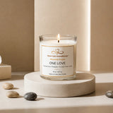 One Love soy candle by Nurse Calm Aromatherapy™ displayed on a neutral cream pedestal with soft spa-inspired décor and warm natural lighting. Clear glass candle with lit flame featuring tropical citrus, pineapple, coconut, rum, and sugar fragrance notes.
