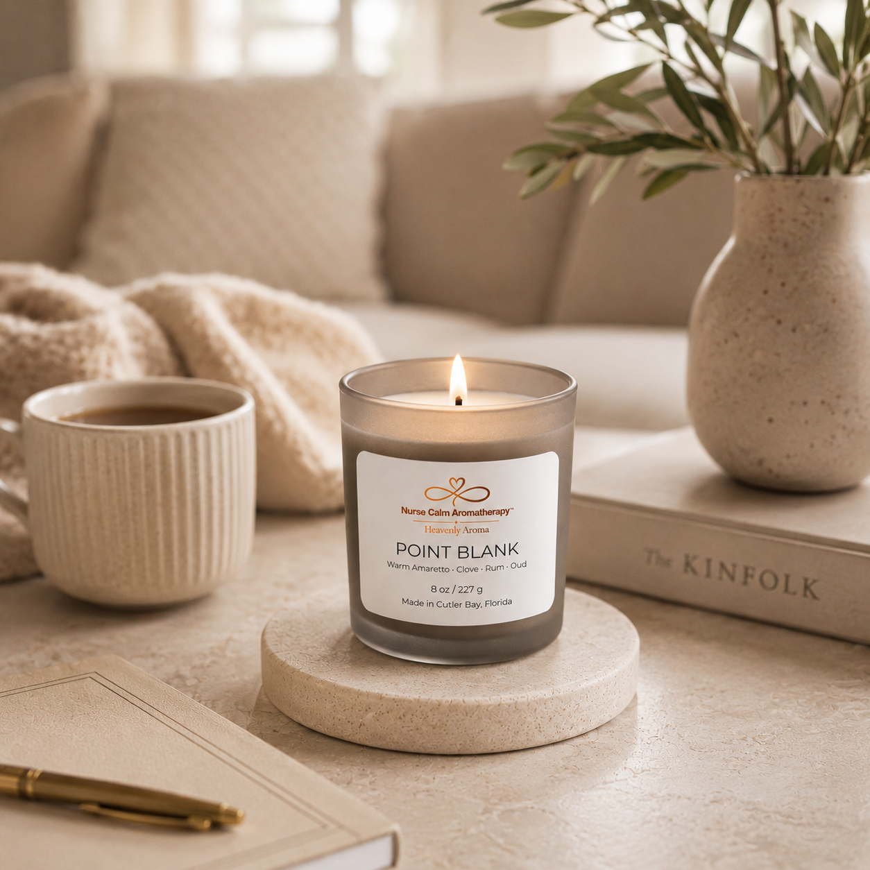Candle with a label on a coaster next to a cup of coffee and a vase with plants in a cozy living room setting.