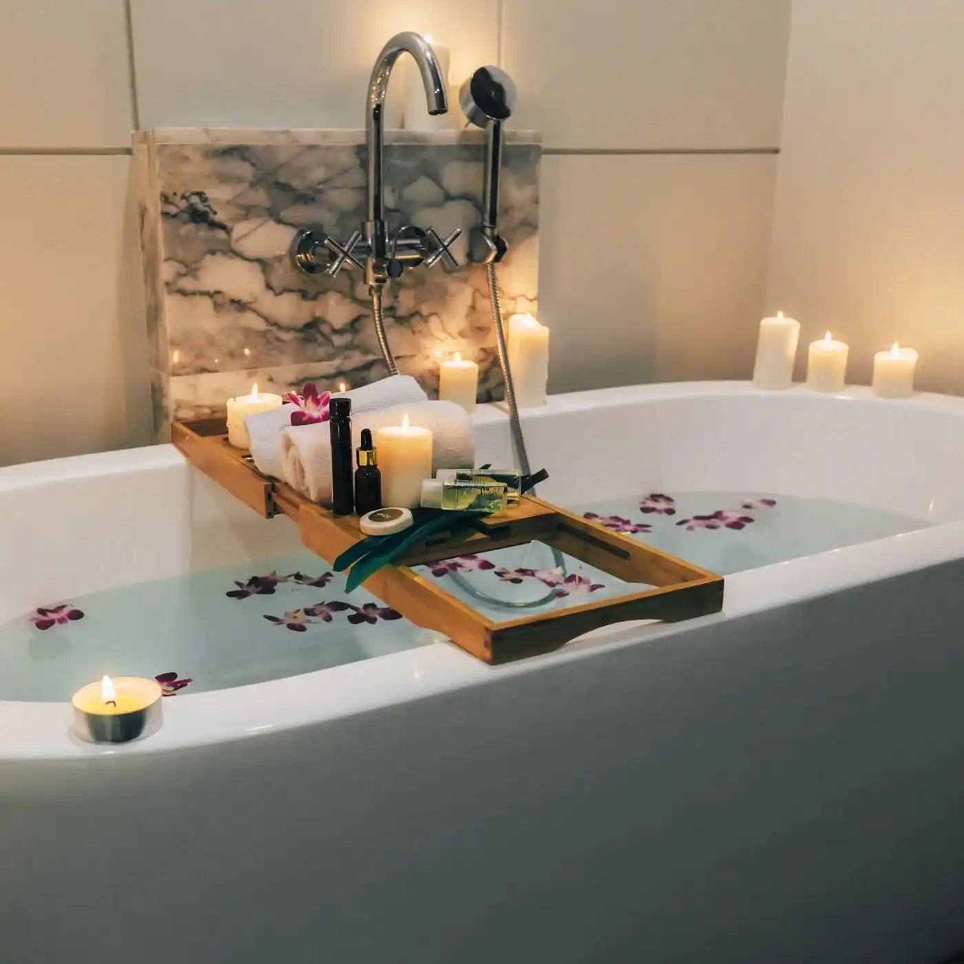 A luxurious white bathtub filled with water and rose petals, featuring a wooden caddy with bath products and candles.